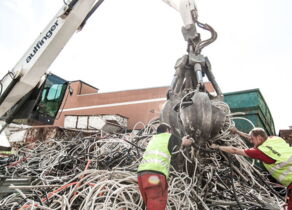 Bagger hebt Kabel aus Kabelhaufen, zwei Mitarbeiter helfen